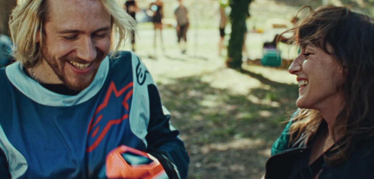 Bastien Bouillon i Juliette Armanet w filmie "Przepis na szczęście" Dwoje ludzi uśmiecha się do siebie na zewnątrz w parku lub lesie. Mężczyzna ubrany jest w strój sportowy, trzyma pomarańczowy przedmiot. W tle widocznych jest kilka nieostrych postaci i drzewa.