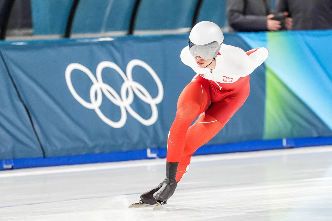Łyżwiarz szybki w stroju reprezentacji Polski dynamicznie pokonuje zakręt toru lodowego. W tle widoczny jest symbol olimpijski.