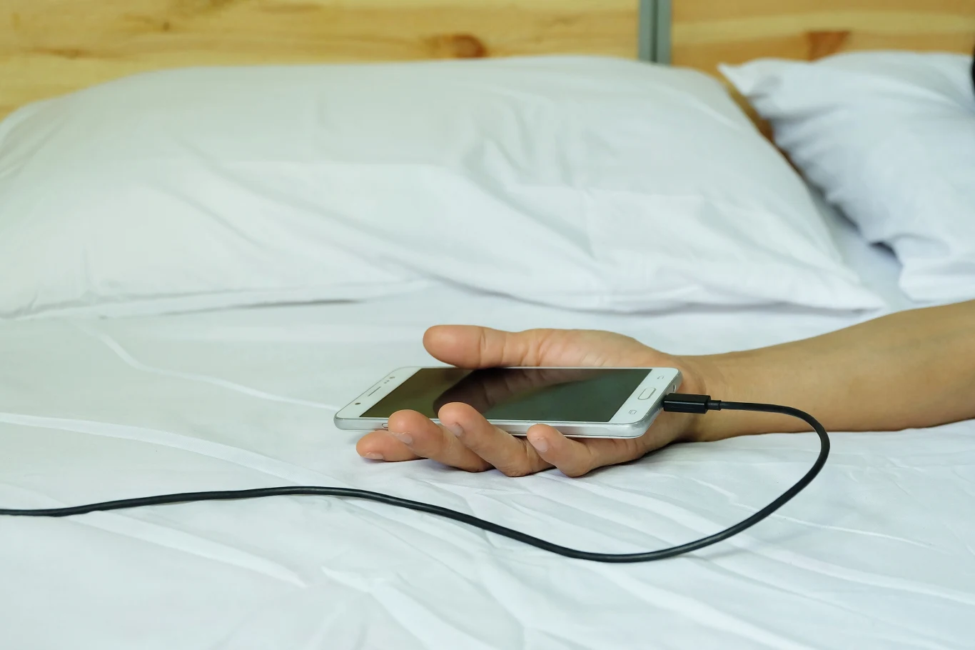 Nigdy nie zasypiaj w pobliżu ładującego się telefonu. Umieść urządzenie z dala od łóżka Ręka leżącej osoby trzymająca smartfon podłączony do ładowarki, telefon leży na białej pościeli, przewód ładowarki widoczny na pierwszym planie.