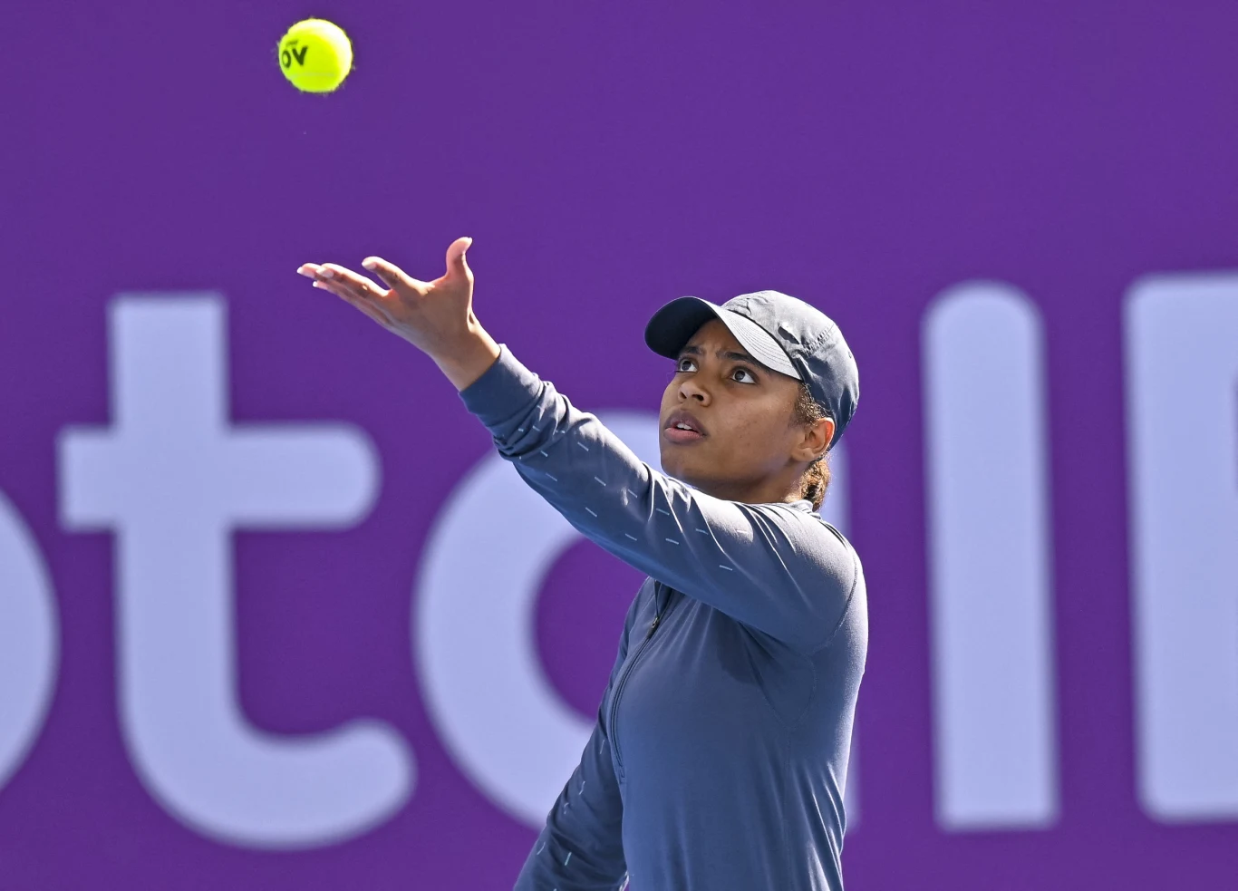 Mubaraka Al-Naimi Tenisistka w sportowym stroju w trakcie serwowania piłki na tle fioletowej ściany z logo. Skoncentrowana, prawa ręka wyciągnięta w górę, piłka tenisowa unosząca się nad dłonią.