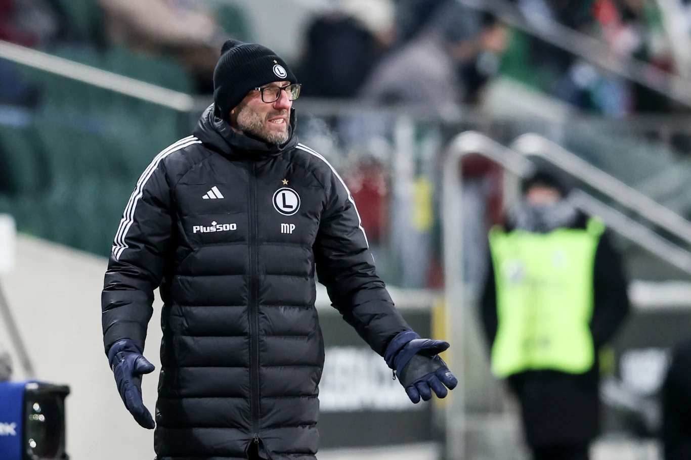 Marek Papszun Trener drużyny piłkarskiej w zimowej kurtce i czapce gestykuluje przy linii bocznej stadionu, w tle rozmazana postać stewarda oraz niewyraźna publiczność na trybunach.