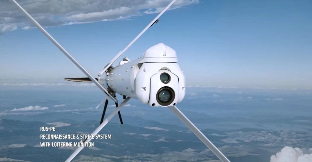 Bezzałogowy dron wojskowy z kamerą, zawieszony w powietrzu wysoko nad chmurami, wyposażony w skrzydła w układzie krzyżowym. Na tle rozległego krajobrazu widać niebieskie niebo oraz delikatne warstwy chmur.