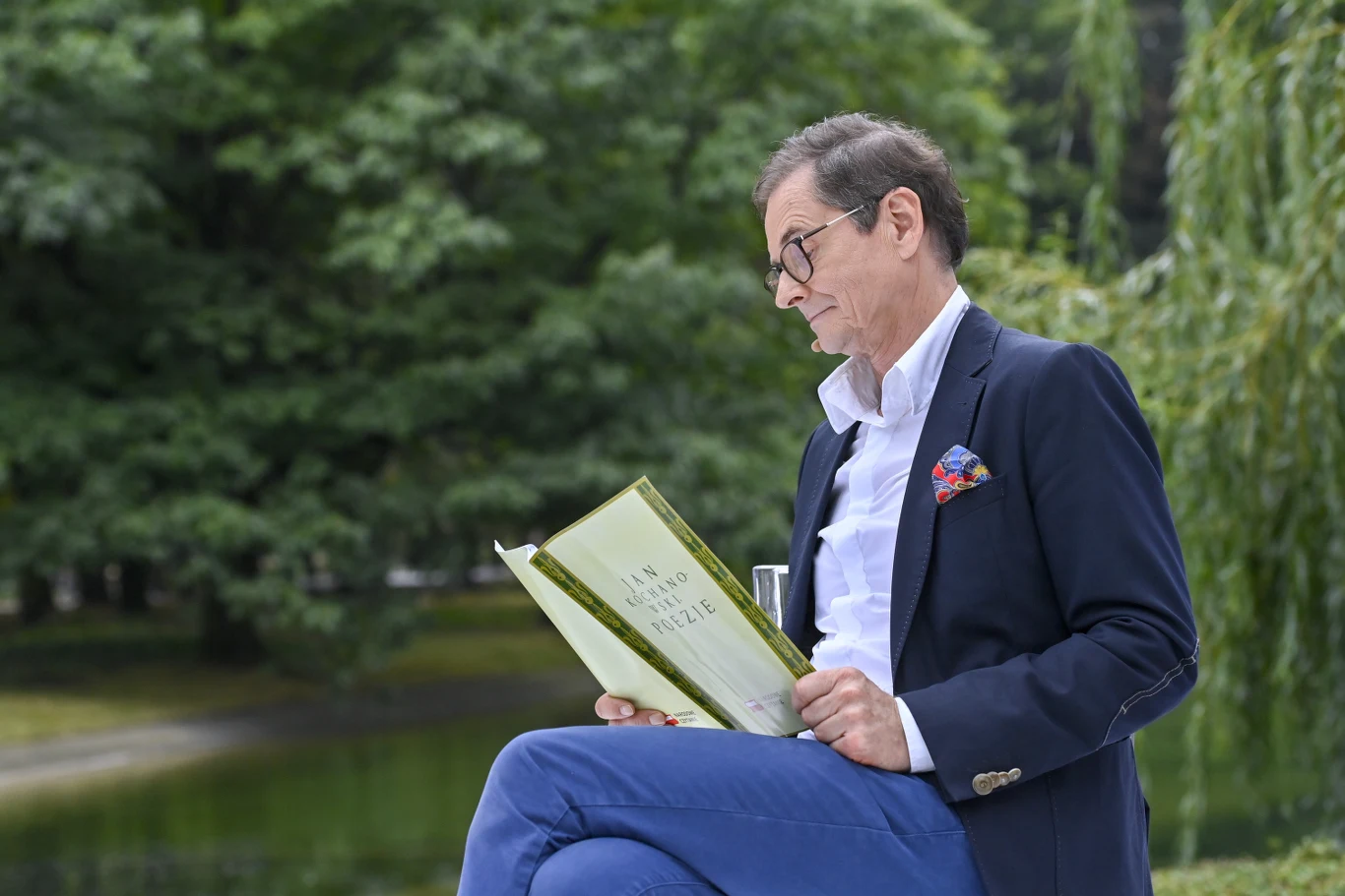 Mężczyzna w okularach, ubrany elegancko w granatową marynarkę i niebieskie spodnie, siedzi nad wodą czytając książkę, w tle zielone drzewa i spokojny park.