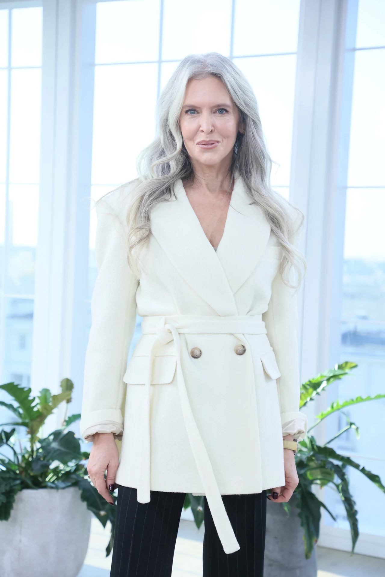 Kobieta o długich, siwych włosach ubrana w elegancką, białą marynarkę przewiązaną paskiem, stoi na tle dużych okien wpuszczających dużo światła. W tle rośliny doniczkowe w jasnym, nowoczesnym wnętrzu.