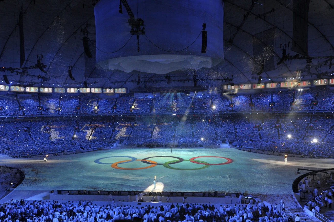 Ceremonia otwarcia zimowych igrzysk 2010 na BC Place Stadium Stadion wypełniony widownią podczas uroczystości z okazji igrzysk olimpijskich, na środku areny widoczny duży symbol olimpijski w postaci pięciu splecionych kół, całość oświetlona niebieskim światłem.
