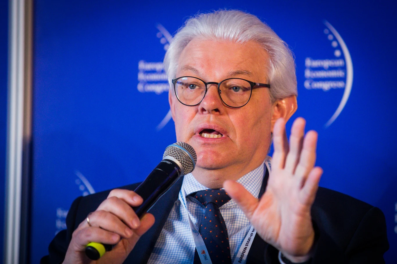 Mężczyzna w okularach z siwymi włosami przemawia do mikrofonu, wykonując gest ręką, na tle niebieskiej ścianki z napisem Kongresu Ekonomicznego.