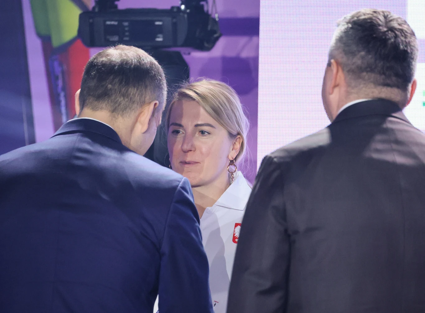 Natalia Czerwonka Trzy osoby w eleganckich ubraniach prowadzą rozmowę w zamkniętym pomieszczeniu, w tle widoczna kamera i rozmyte elementy scenografii, na pierwszym planie kobieta z przypinką w barwach narodowych.