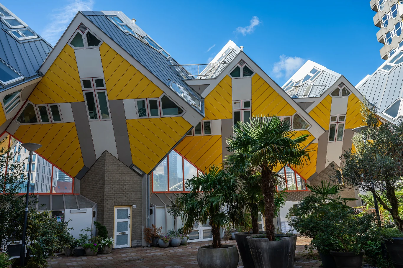 Nowoczesne budynki o charakterystycznej żółtej, skośnej konstrukcji z dużymi oknami, otoczone roślinami doniczkowymi na dziedzińcu, pod błękitnym niebem.