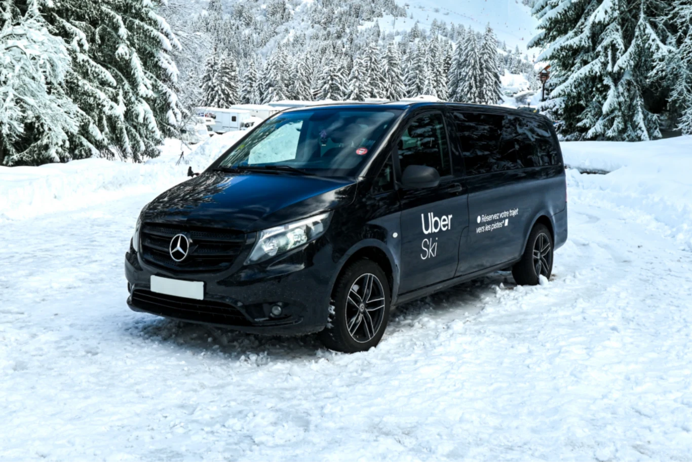 Czarny samochód dostawczy marki Mercedes z oznaczeniem Uber Ski zaparkowany na ośnieżonej drodze, w otoczeniu zaśnieżonych drzew i górskiego krajobrazu.