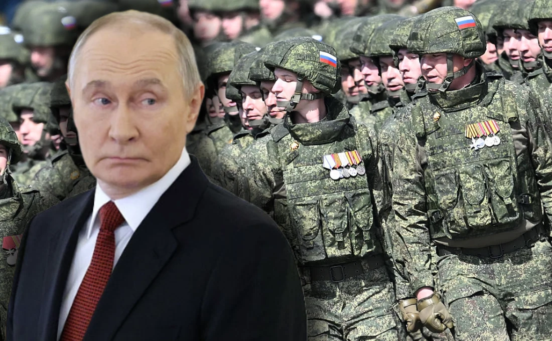 Widoczny mężczyzna w garniturze na pierwszym planie oraz oddział żołnierzy w mundurach wojskowych z odznaczeniami i rosyjskimi flagami na rękawie maszerujący w szyku.