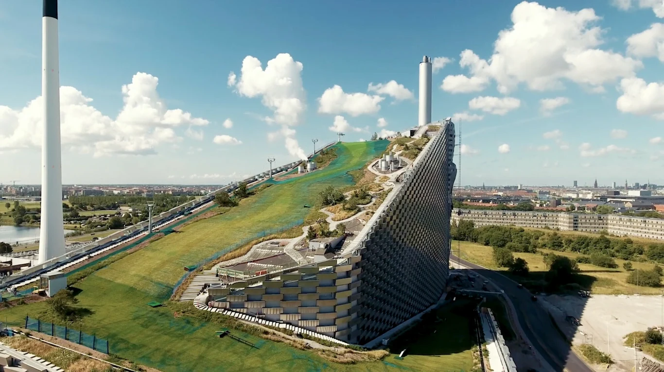 Nowoczesny budynek z pochylonym dachem, na którym znajdują się trasy do zjazdów i tereny zielone. Konstrukcja otoczona jest przez otwartą przestrzeń, w oddali widoczna panorama miasta i wysoki komin elektrociepłowni.