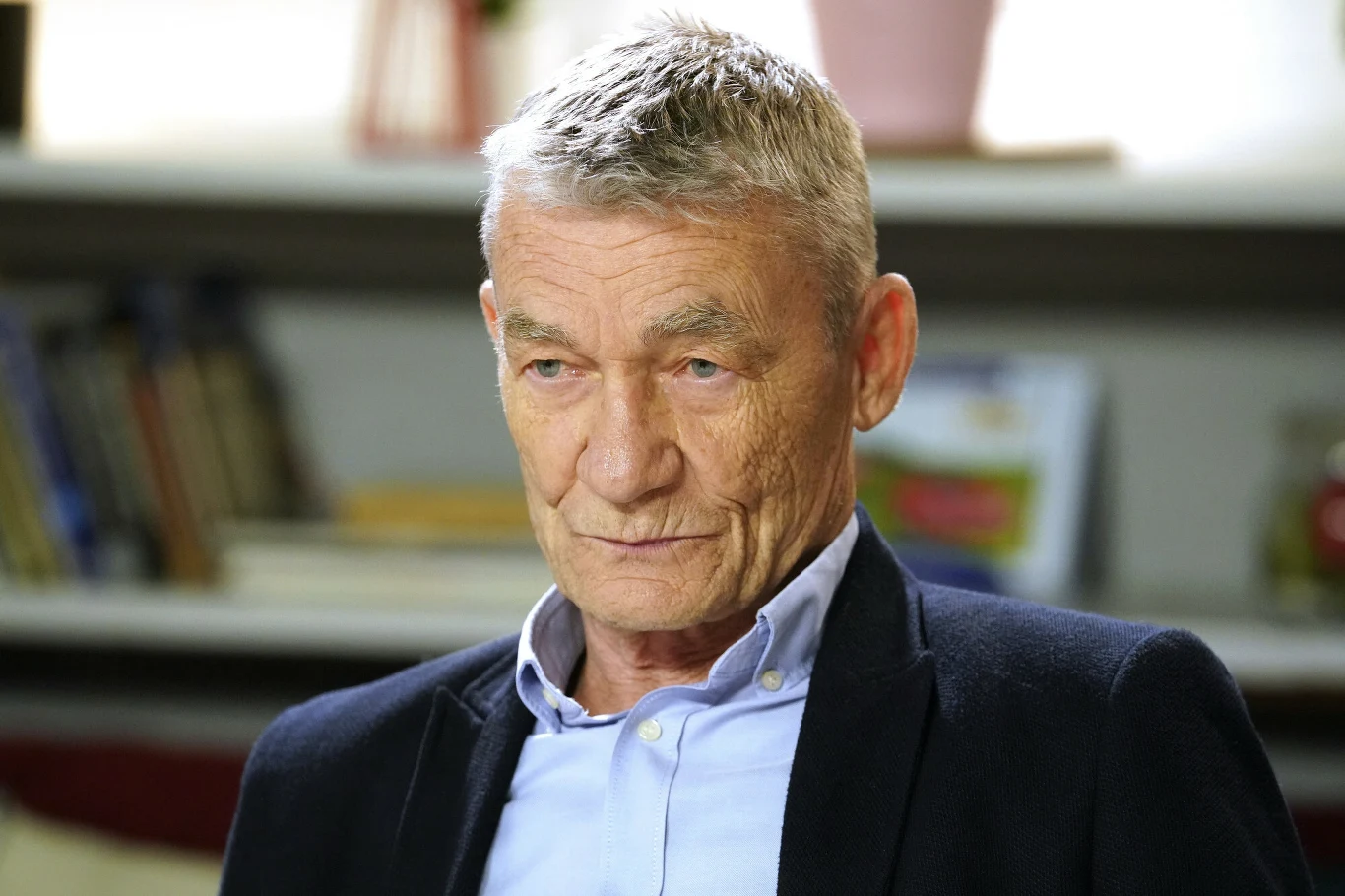 Krzysztof Kiersznowski Starszy mężczyzna w marynarce i jasnej koszuli, siedzący w pomieszczeniu z półkami na książki w tle, wyraz twarzy poważny, włosy krótkie i siwe.