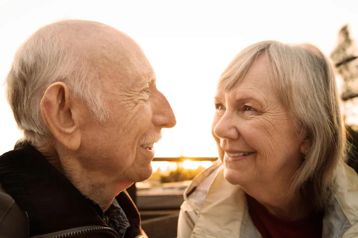 Dwoje starszych ludzi patrzących na siebie z uśmiechem, twarze oświetlone ciepłym światłem zachodzącego słońca, atmosfera bliskości i szczęścia.