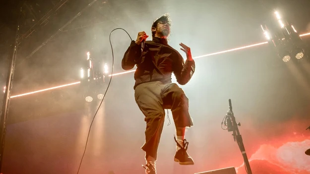 Wokalista skaczący na scenie podczas koncertu, trzymający mikrofon w dłoni, otoczony efektownym oświetleniem i dynamicznym tłem.