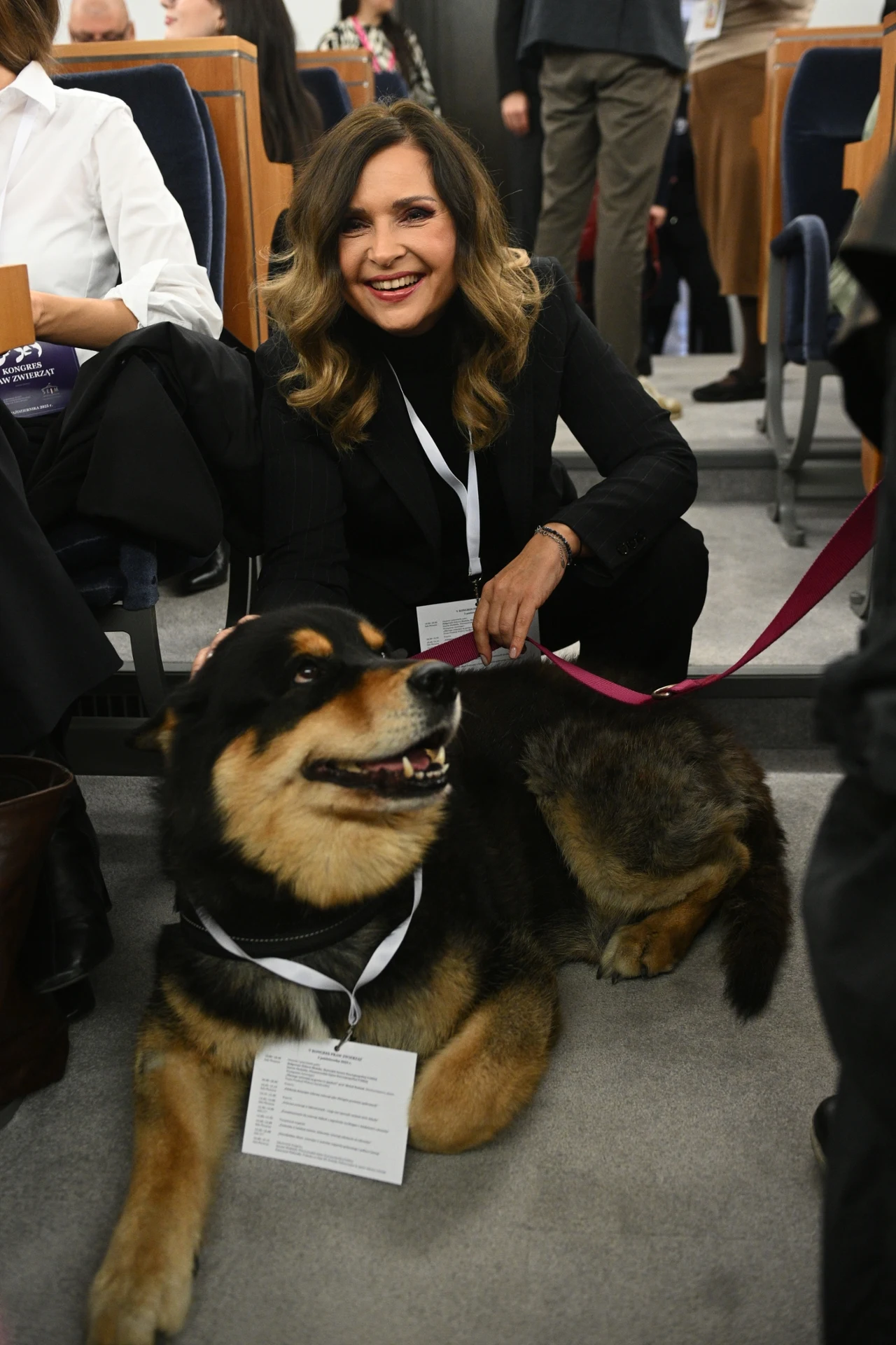 Uśmiechnięta kobieta w czarnym stroju kuca obok dużego, przyjaznego psa z identyfikatorem na szyi. W tle widać inne osoby siedzące na sali konferencyjnej.