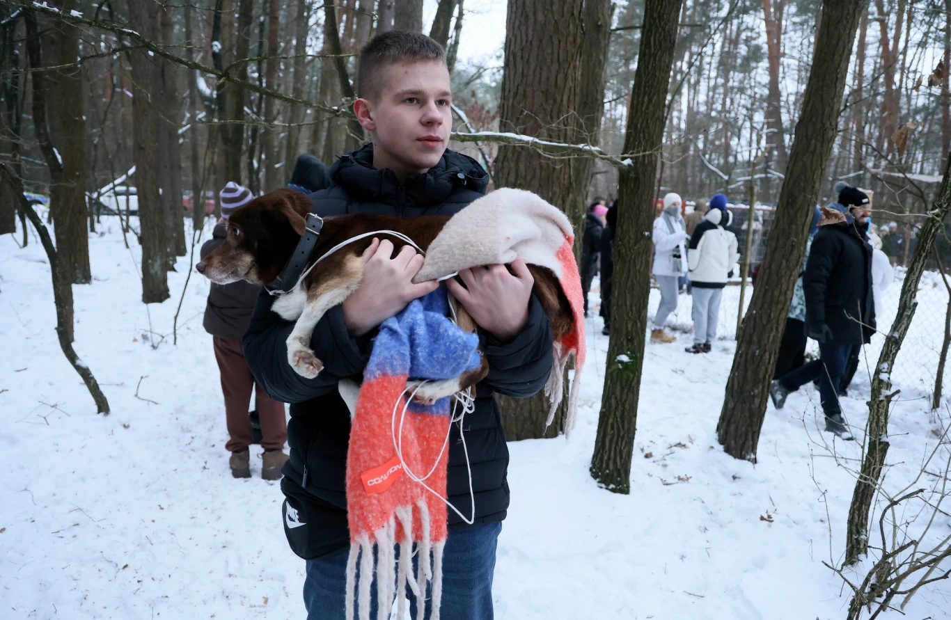 Młody mężczyzna trzyma na rękach psa okrytego kocem, wokół niego las w zimowej scenerii oraz grupa ludzi w tle, wszyscy ubrani ciepło na zimę.
