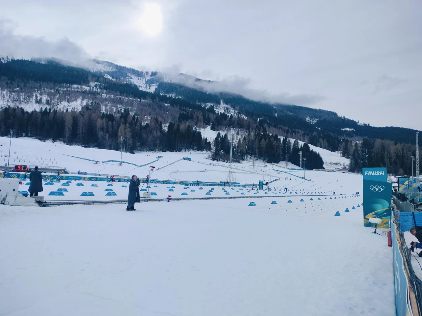 Zaśnieżone pole narciarskie z wyznaczonymi trasami oznaczonymi niebieskimi pachołkami, w tle widoczne góry oraz fragment lasu, po prawej stronie meta oznaczona banerem olimpijskim.