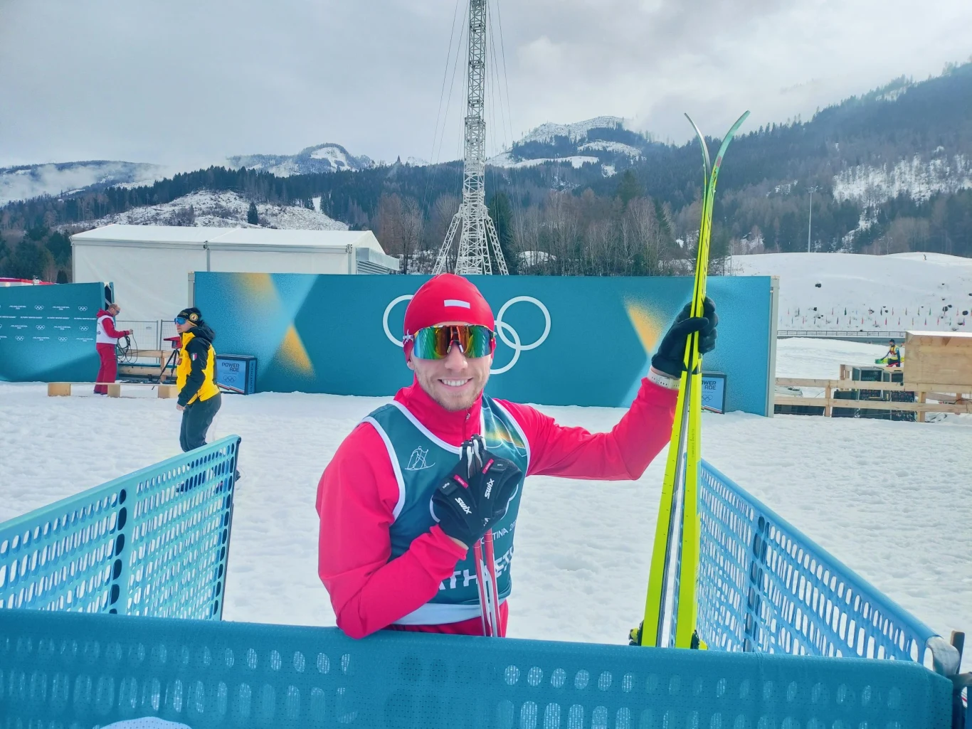 Uśmiechnięty narciarz w stroju sportowym trzymający narty, stojący przy niebieskiej barierce na tle zaśnieżonych gór i logotypu olimpijskiego. W tle widać inne osoby i zimową infrastrukturę sportową.