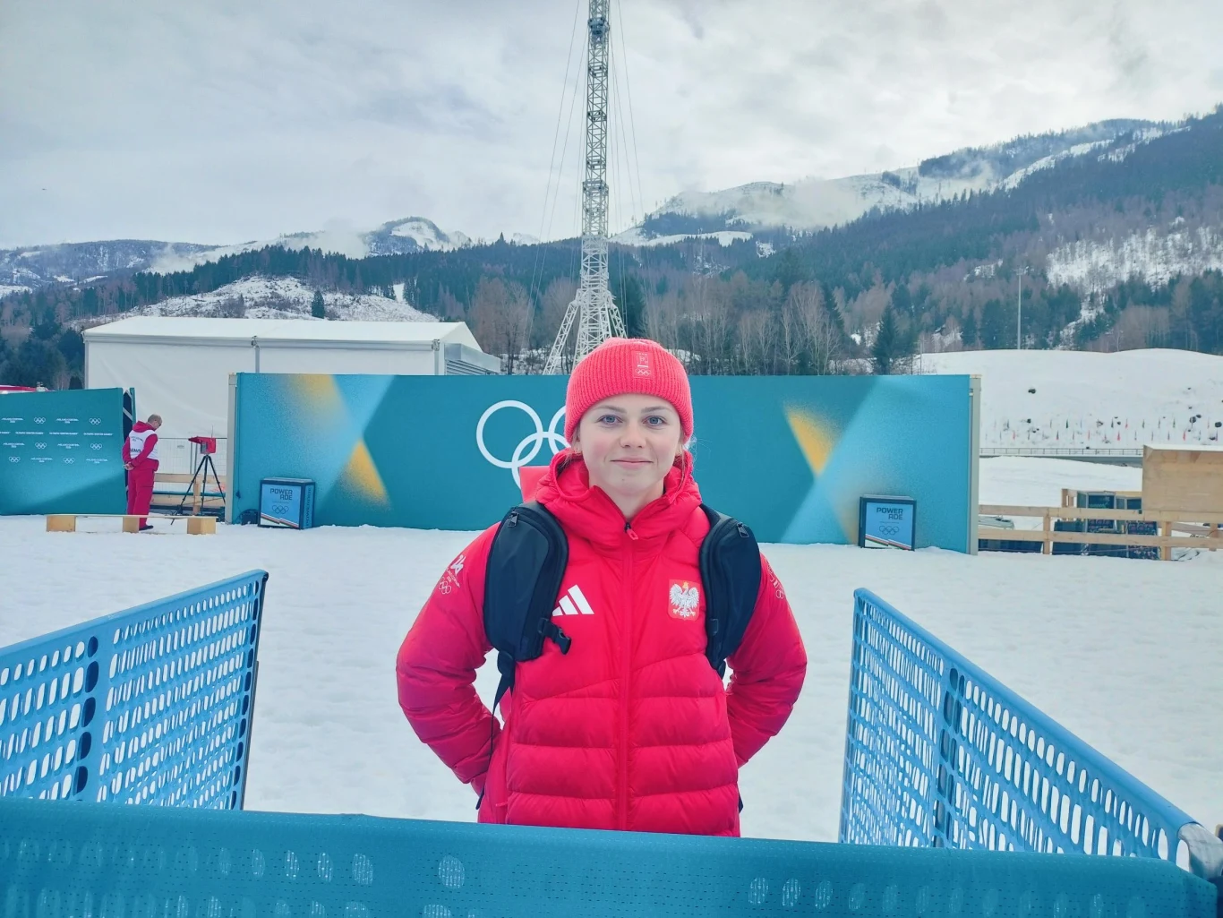 Osoba ubrana w czerwony zimowy strój reprezentująca Polskę na tle śnieżnego krajobrazu, gór i obiektów sportowych z symbolami olimpijskimi, otoczona metalowym ogrodzeniem.