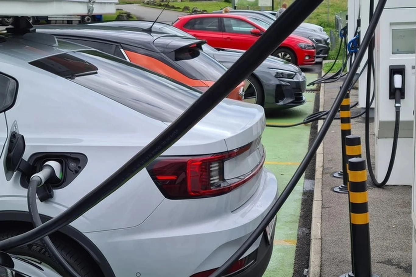 Rząd Kanady zdecydował o miliardowych dopłatach do elektryków Kilka nowoczesnych samochodów elektrycznych zaparkowanych przy stacji ładowania z podłączonymi kablami. W tle widoczny zielony pas i asfalt, a także słupki zabezpieczające oraz ekran terminala ładowania.