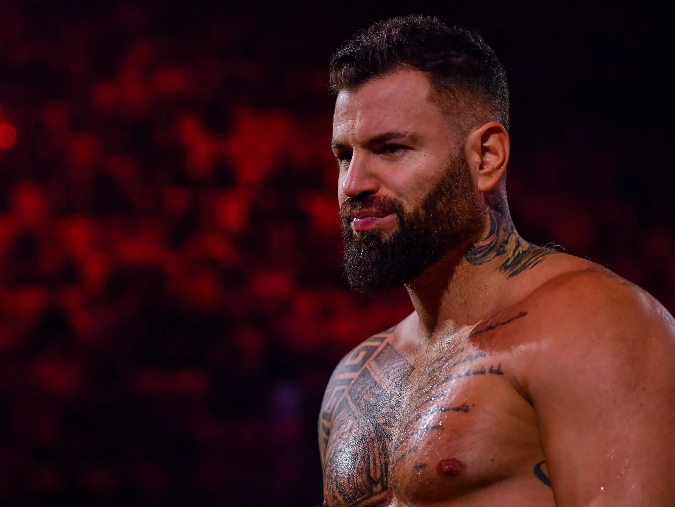 Wojciech Gola Umięśniony mężczyzna z brodą i licznymi tatuażami na górnej części ciała stoi na tle rozmytej, ciemnoczerwonej publiczności, patrząc zdecydowanie w bok.