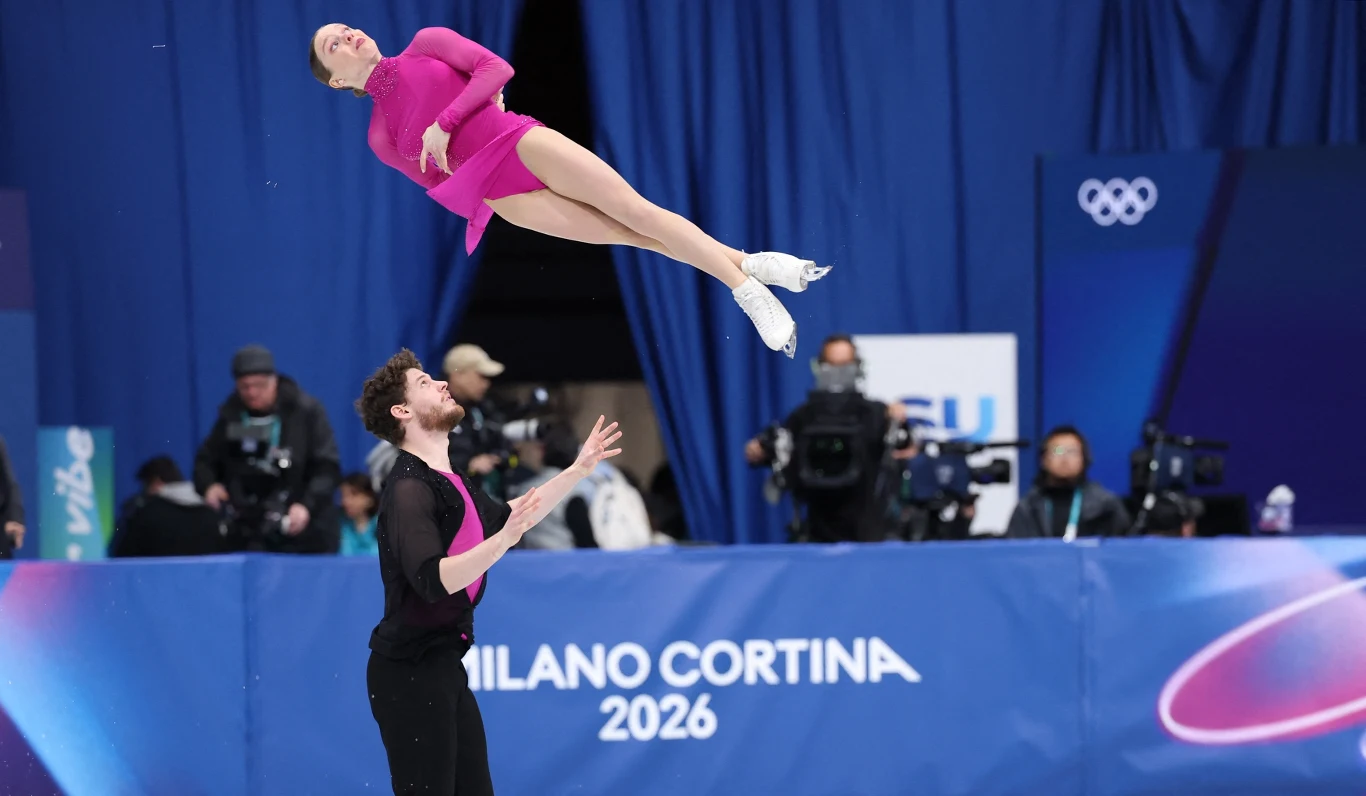 Duet łyżwiarzy figurowych wykonuje skomplikowany podnoszenie, kobieta w różowym stroju leci wysoko nad partnerem, w tle błękitne dekoracje, logo igrzysk Olimpijskich oraz obserwujący fotoreporterzy.