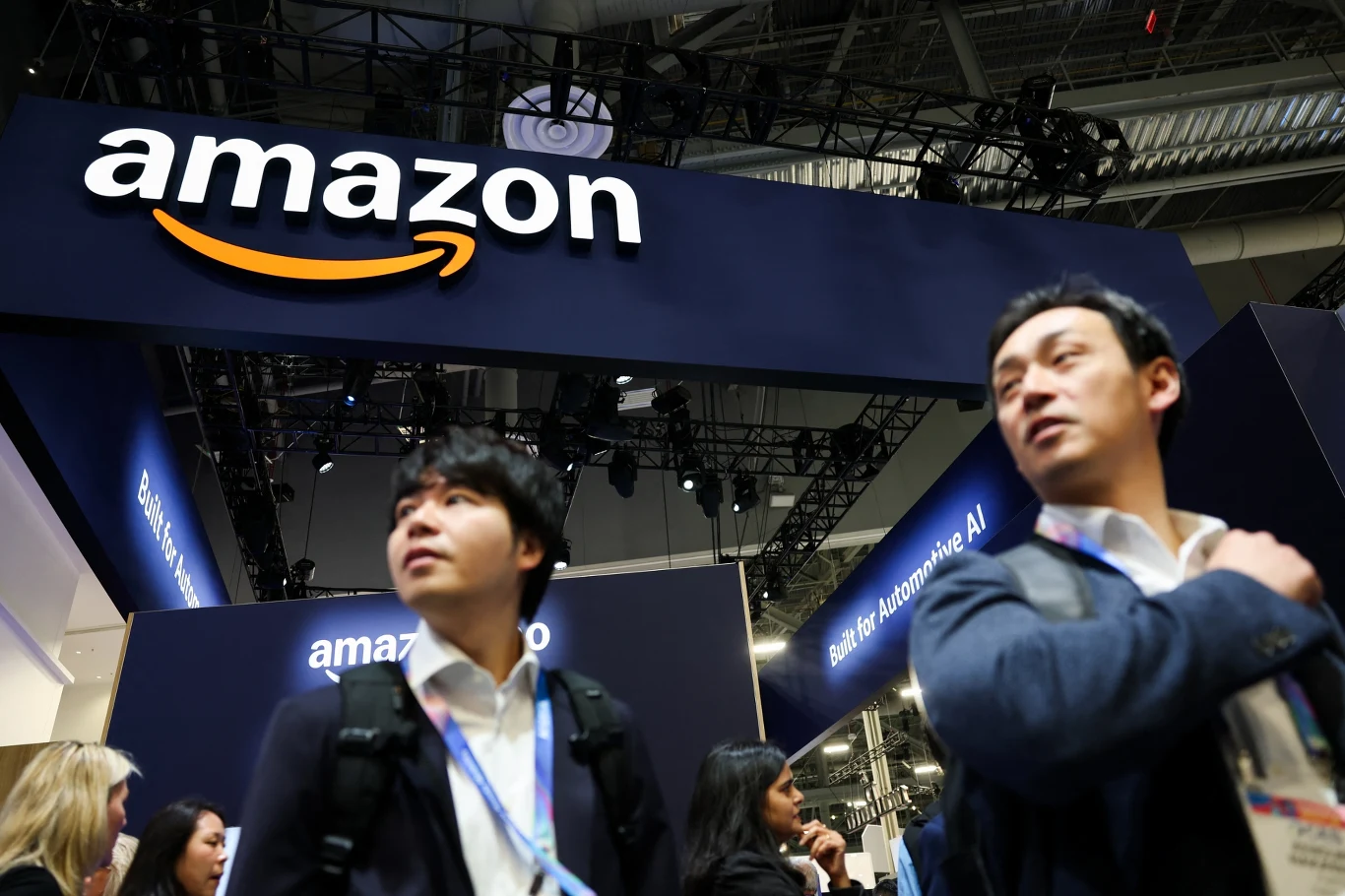 Amazon dołączył do koncernów zapowiadających liczone w setkach miliardów dolarów wydatki na rozwój AI. Akcje Big Techów spadają - inwestorzy boją się pęknięcia "bańki AI" Dwóch mężczyzn w garniturach z identyfikatorami na szyi, stojących pod dużym szyldem z logo Amazon na tle targów lub konferencji. W tle widoczne są elementy stoiska oraz inni uczestnicy wydarzenia.