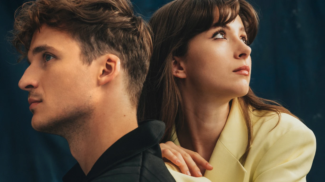 Dwoje młodych dorosłych patrzących w przeciwnych kierunkach na ciemnym tle, kobieta w żółtym żakiecie trzyma rękę na ramieniu mężczyzny.