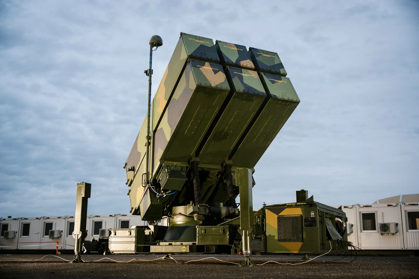Wyrzutnia rakiet przeciwlotniczych ustawiona na twardym podłożu, w tle widoczne jasne baraki biurowe pod zachmurzonym niebem.