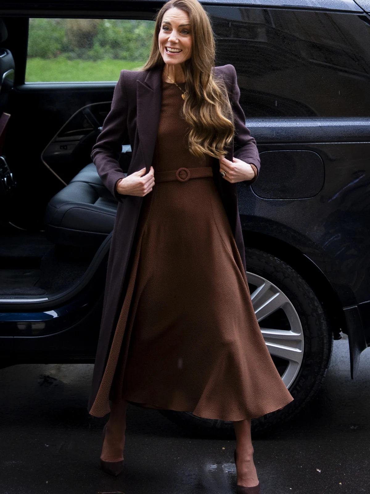 Kobieta z długimi, rozpuszczonymi włosami w eleganckiej, brązowej sukience i ciemnym płaszczu stoi obok czarnego samochodu z uśmiechem na twarzy.