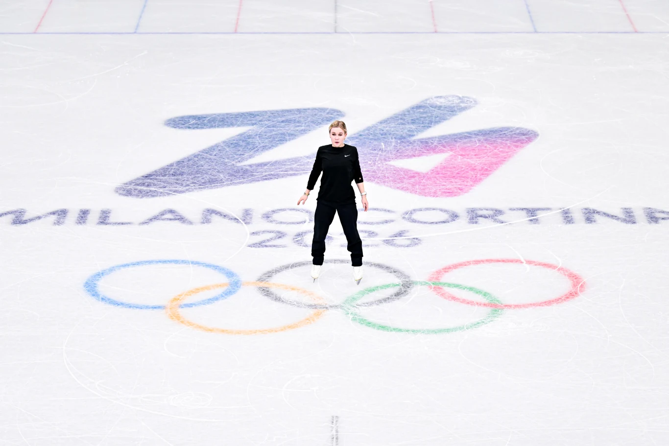 Łyżwiarz figurowy stoi na środku lodowiska z dużym logo Z4 oraz napisem Milano Cortina 2026 i symbolami olimpijskimi w tle.
