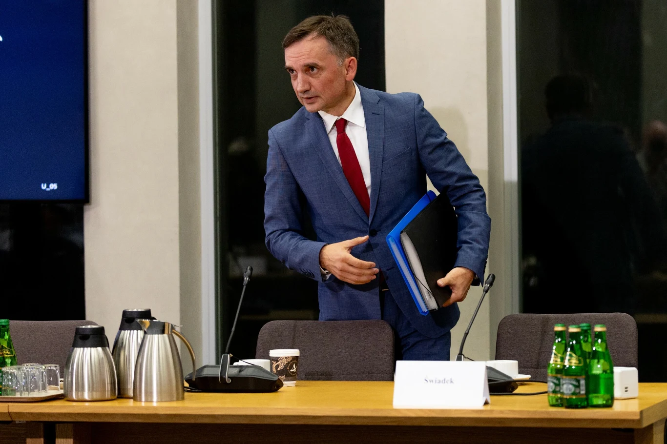 Sąd w Warszawie zdecydował o areszcie dla Zbigniewa Ziobry, za politykiem wystawiony został list gończy Mężczyzna w garniturze z czerwonym krawatem stoi przy stole konferencyjnym, trzymając segregator i dokumenty. Na stole stoją butelki z wodą, metalowe termosy i filiżanka. W tle widoczne są duże okna i tabliczka z napisem.