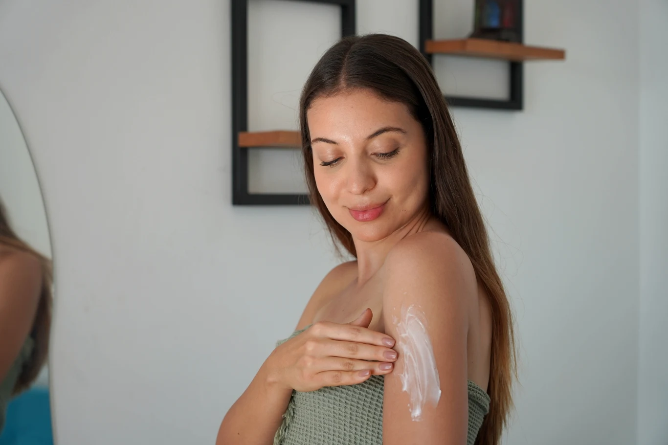 Młoda kobieta nakłada krem na ramię, stojąc w jasnym pomieszczeniu z nowoczesnymi półkami na ścianie.