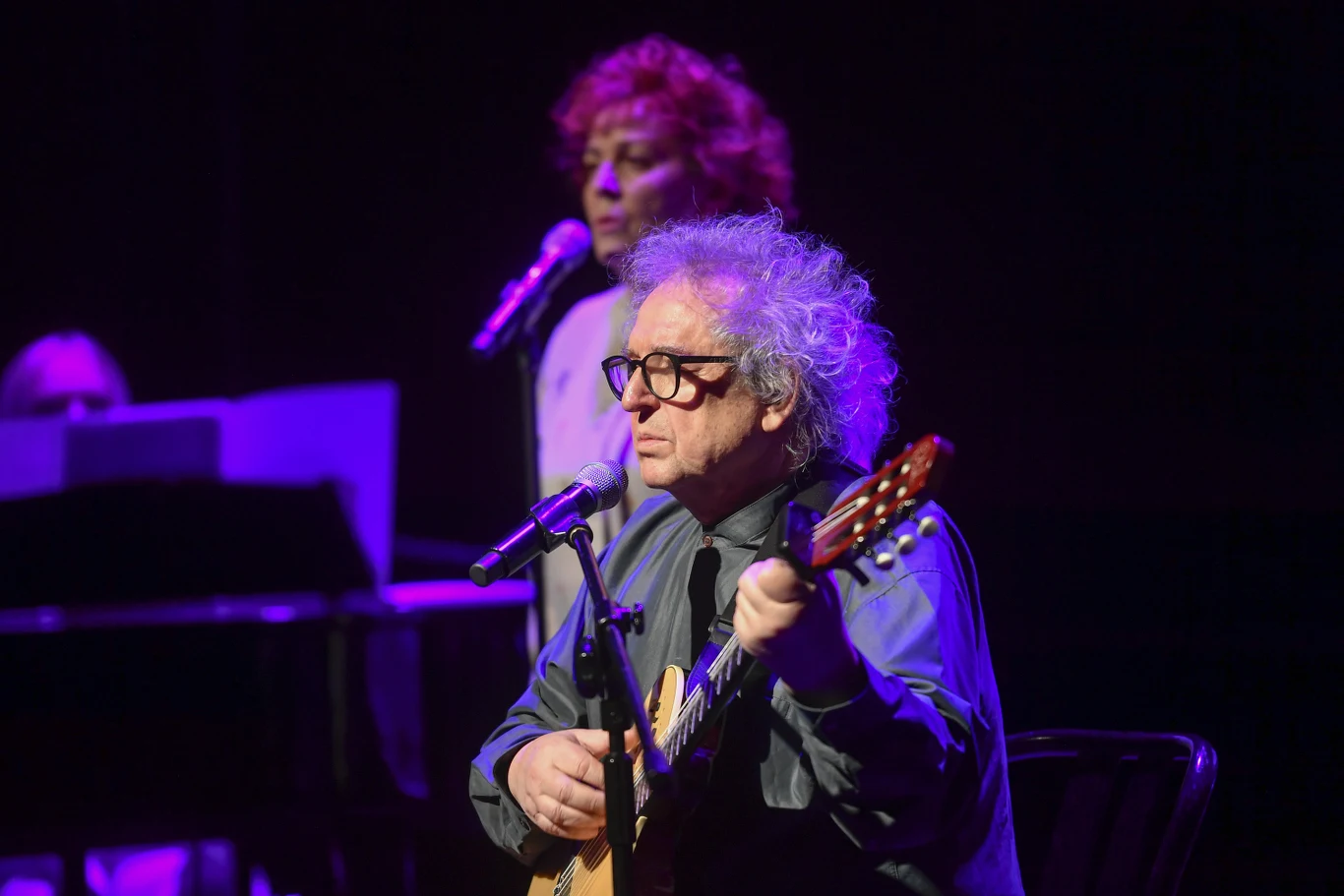 Tadeusz Woźniak Mężczyzna z siwymi, kręconymi włosami i czarnymi okularami śpiewa i gra na gitarze na scenie, w tle druga osoba przy mikrofonie, scena oświetlona fioletowym światłem.