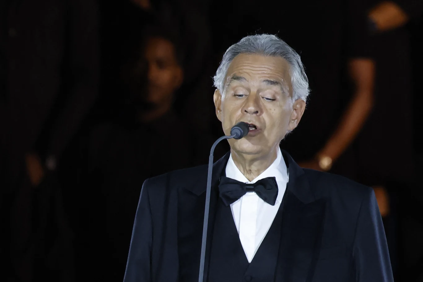 Andrea Bocelli Elegancko ubrany mężczyzna w smokingu z muchą śpiewa do mikrofonu, na ciemnym tle widoczne zamazane sylwetki innych osób.