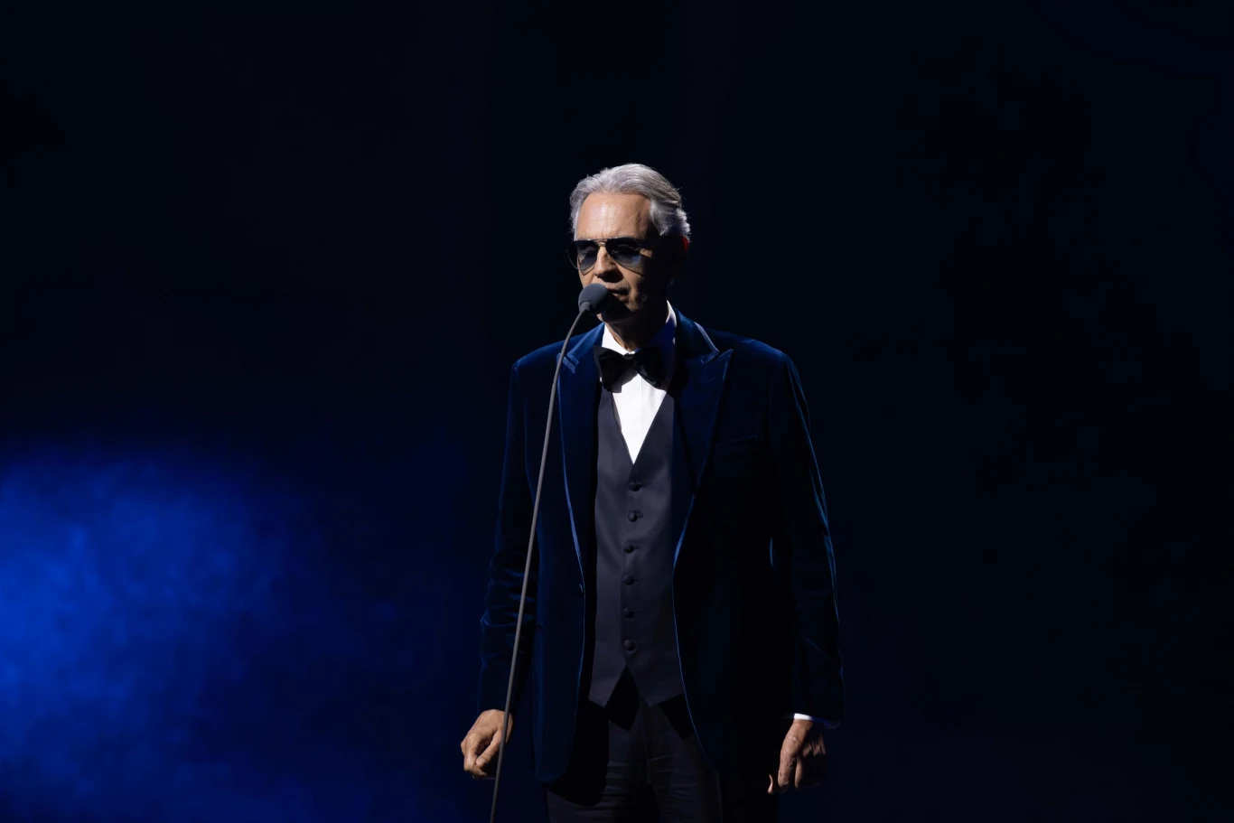 Andrea Bocelli Mężczyzna w eleganckim garniturze, stojący na scenie z mikrofonem, oświetlony światłem z boku, na ciemnym tle.