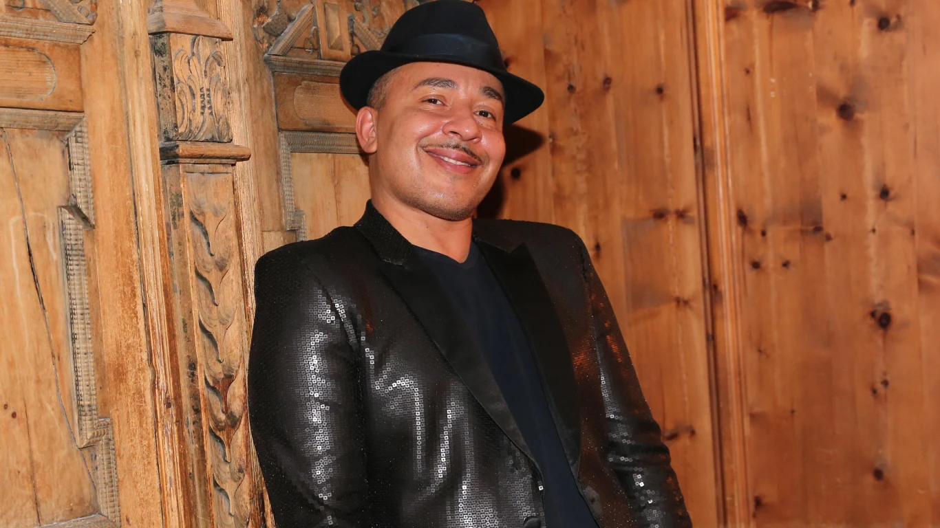 Lou Bega Mężczyzna w czarnym błyszczącym garniturze i ciemnym kapeluszu stoi oparty o drewnianą dekorację ścienną, uśmiechając się szeroko. Za nim znajduje się drewniana ściana o ciepłym odcieniu.