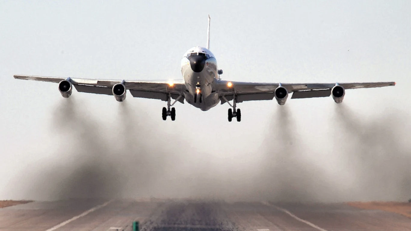 Duży samolot pasażerski w trakcie startu z pasa startowego, silniki wydzielają ciemny dym, wokół jasne, pustynne otoczenie, a maszyna unosi się stopniowo nad ziemią.