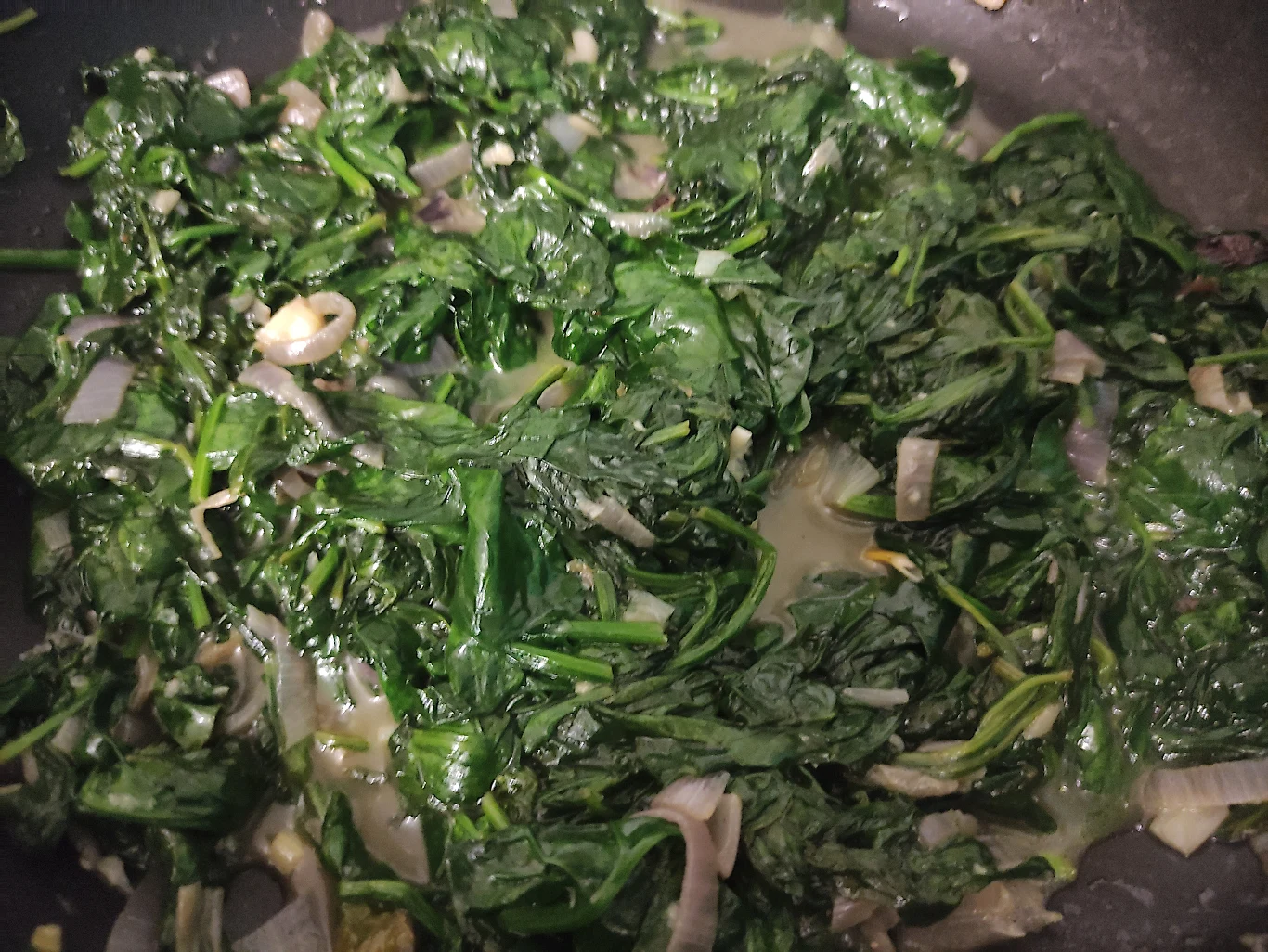 Szpian duś po przykryciem Liście szpinaku podsmażane na patelni z dodatkiem pokrojonej cebuli i czosnku. Zielone liście są miękkie i lekko wilgotne, widoczny jest sos powstały podczas smażenia.