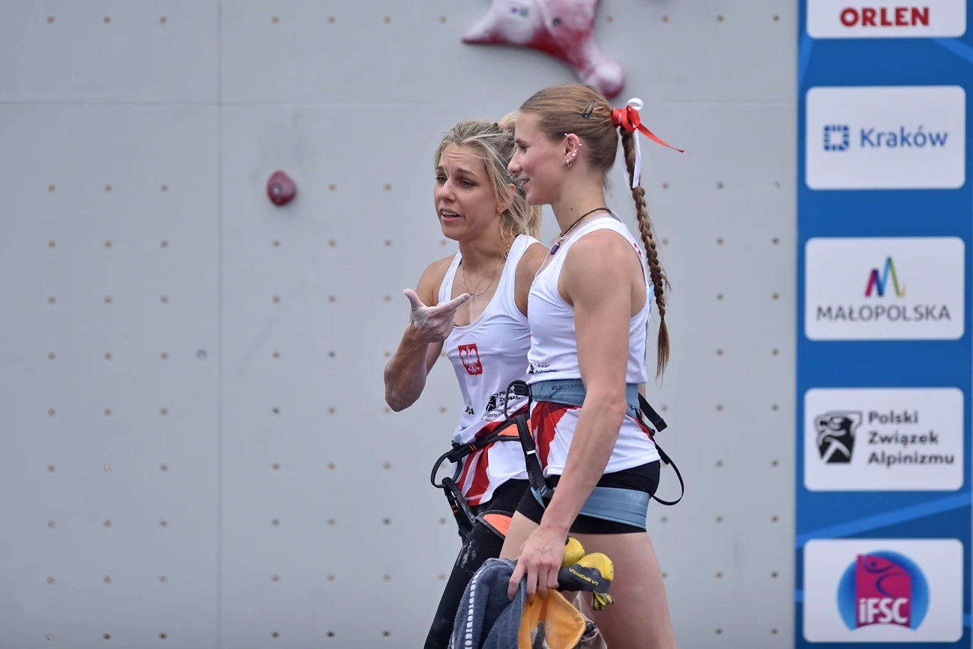Aleksandra Mirosław i Natalia Kałucka Dwie kobiety w sportowych uprzężach wspinaczkowych stoją przy ściance wspinaczkowej, jedna z nich gestykuluje podczas rozmowy, obok widoczna tablica z logotypami sponsorów oraz instytucji związanych ze wspinaczką.