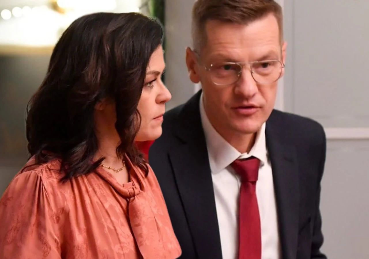 Katarzyna Cichopek i Marcin Mroczek na planie serialu "M jak miłość" Kobieta z ciemnymi włosami w eleganckiej, różowej bluzce i mężczyzna w okularach w garniturze z czerwoną krawatą rozmawiają ze sobą w jasnym pomieszczeniu.