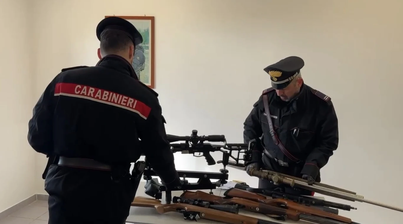 Dwóch funkcjonariuszy Carabinieri stoi przy stole, na którym leży kilka sztuk długiej broni palnej, w tym karabiny oraz lunety celownicze. Mundury są wyraźnie oznaczone napisem jednostki.