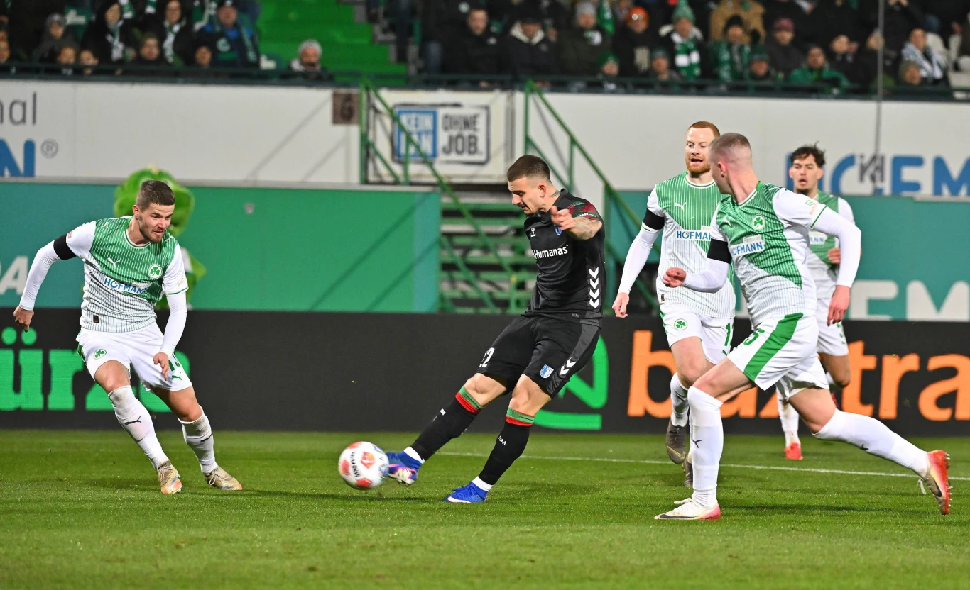 Piłkarz w czarnym stroju próbuje oddać strzał na bramkę otoczony przez czterech zawodników drużyny przeciwnej w biało-zielonych strojach podczas meczu piłkarskiego na stadionie z widocznymi kibicami na trybunach.