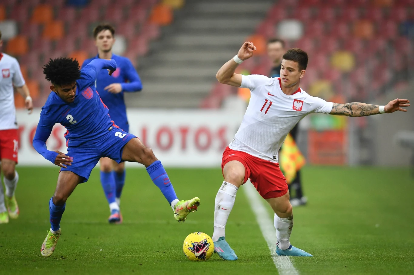 Dwóch piłkarzy walczy o piłkę na boisku, jeden w niebieskim stroju reprezentuje drużynę Anglii, drugi w białym stroju z czerwonymi elementami reprezentuje drużynę Polski, w tle zawodnicy i trybuny stadionu.