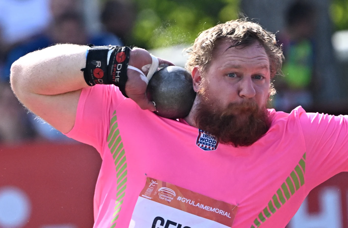 Jordan Geist Mężczyzna w intensywnie różowej koszulce przygotowuje się do pchnięcia kulą, trzymając ciężką kulę przy brodzie, z wyrazem koncentracji na twarzy. W tle rozmyte postaci i elementy stadionu.