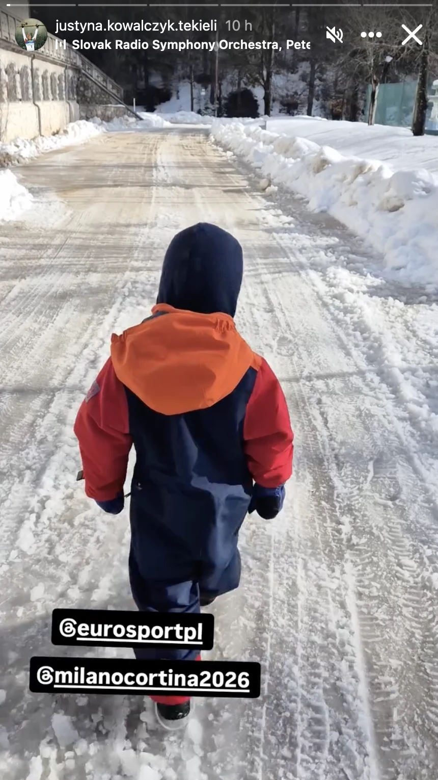 Justynie Kowalczyk-Tekieli w Cortinie d'Ampezzo towarzyszy jej syn Hugo Dziecko w kolorowym kombinezonie zimowym idzie samotnie po zaśnieżonej, oczyszczonej drodze, otoczonej wysokimi zaspami śniegu i drzewami w tle.