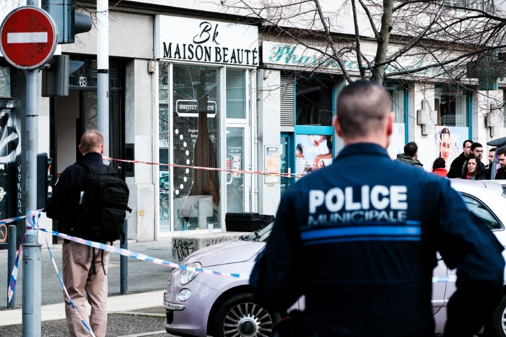 Policjant oraz inne osoby stoją przed budynkiem oznaczonym taśmą policyjną, w tle widoczny salon kosmetyczny oraz grupka ludzi obserwująca sytuację.