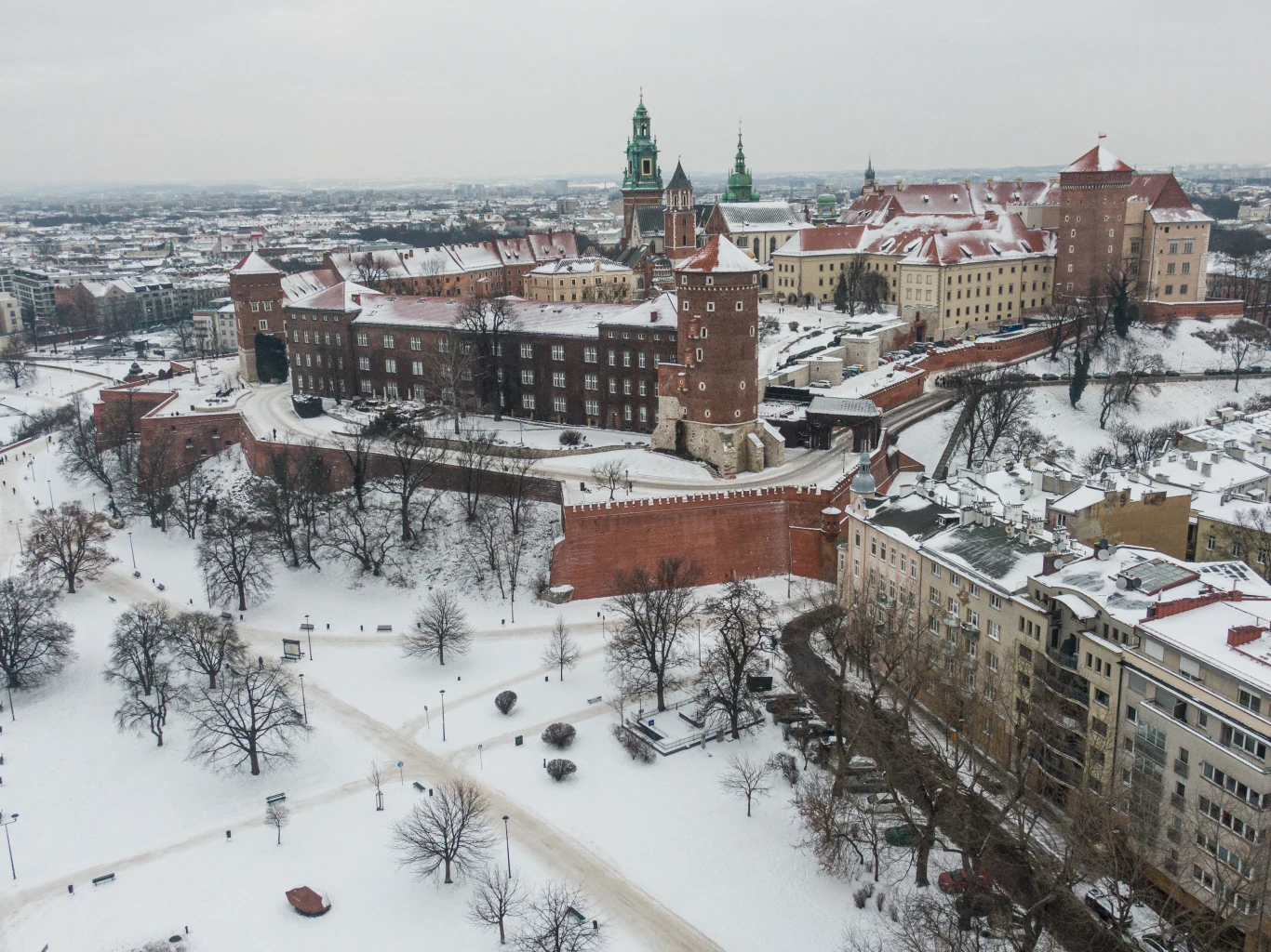 Zamek królewski Wawel otoczony zimową scenerią, pokryty warstwą śniegu, z wyraźnie widocznymi murami obronnymi, wieżami i otaczającymi go ulicami oraz budynkami mieszkalnymi.