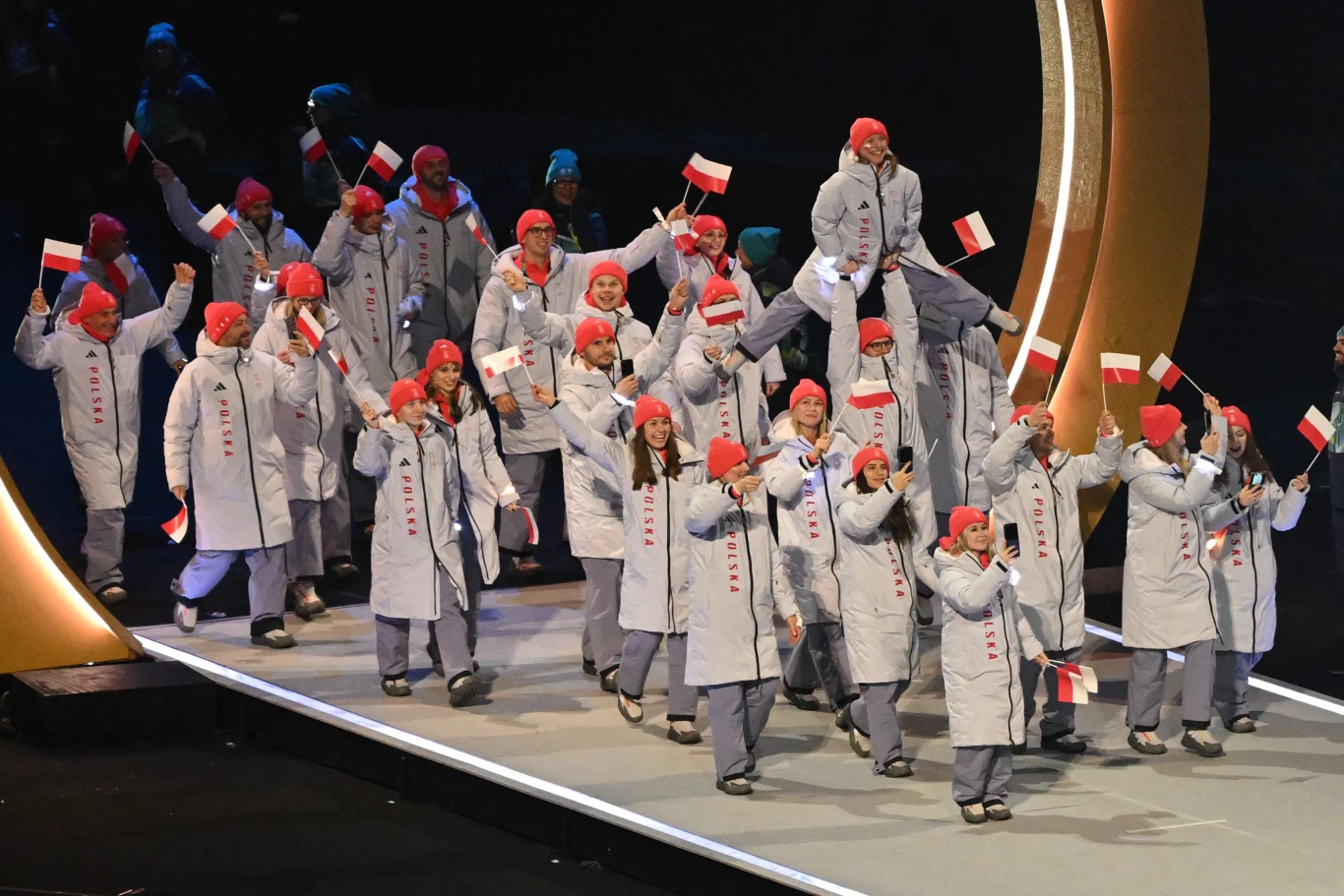 Grupa sportowców ubrana w jednolite, jasnoszare kurtki i czerwone czapki z biało-czerwonymi flagami maszeruje na tle dużego, złotego pierścienia, wyrażając radość i dumę narodową.