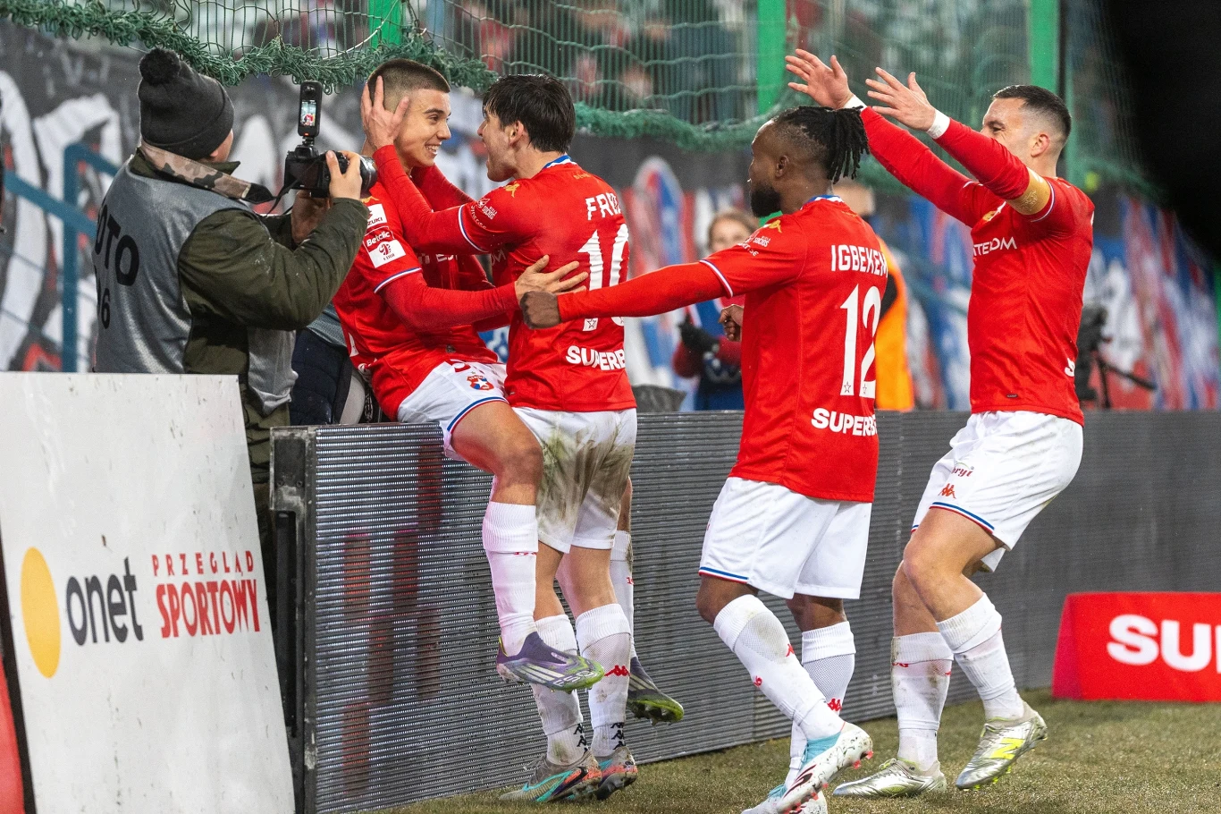 Maciej Kuzienka (z lewej) zdobył dwie bramki dla Wisły Kraków Grupa piłkarzy w czerwonych koszulkach świętuje zdobycie bramki przy bandzie reklamowej, jeden z zawodników wskakuje na bandę i zwraca się do fotografa, pozostali piłkarze unoszą ręce w geście radości.